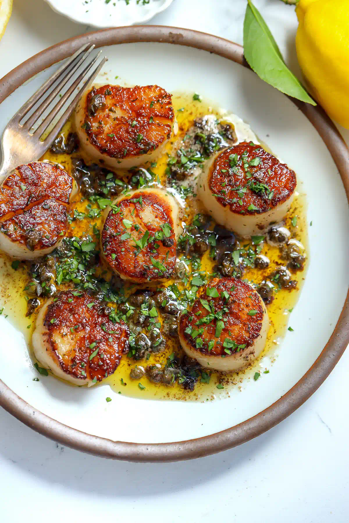 Overhead view of scallops meunière on a white plate with lemon caper butter sauce, fresh herbs, and a whole lemon