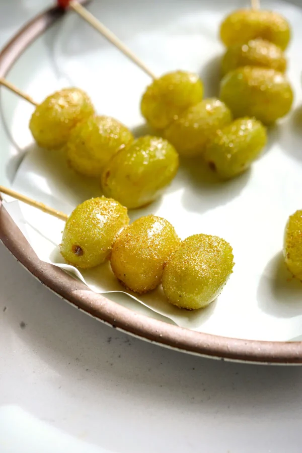 Close-up of gold-sugared champagne grapes on a wooden skewer resting on a speckled cream plate.