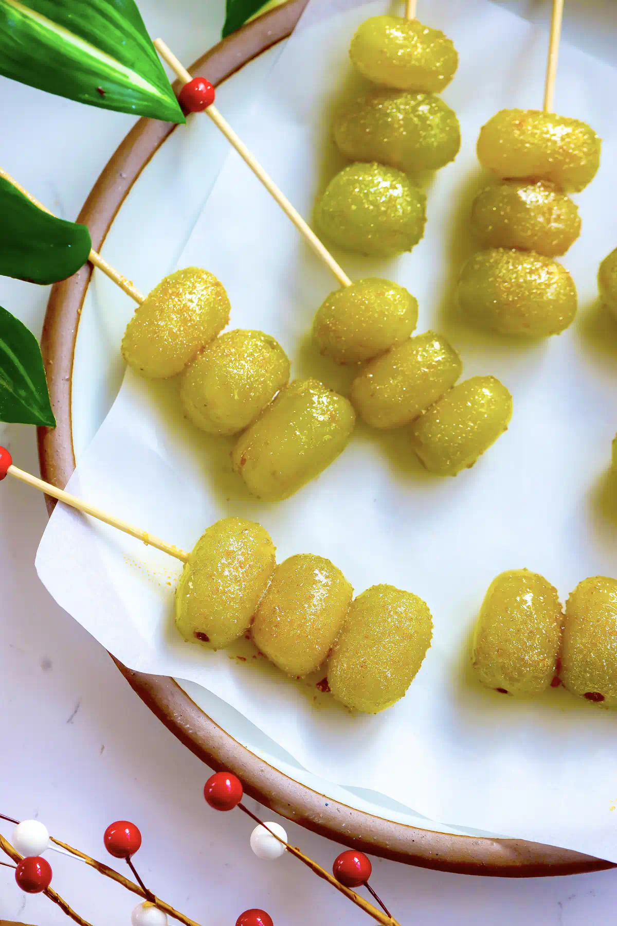 Champagne grapes rolled in gold sugar and threaded onto wooden skewers, plated on parchment with red berries and green leaves