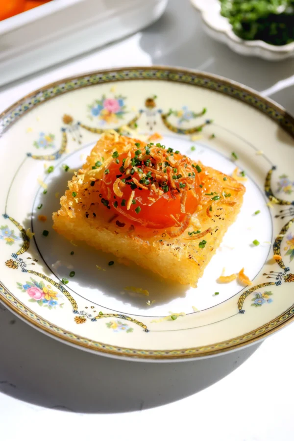 Cured egg yolk crispy rice bite topped with chives and crispy shallots on a decorative plate.