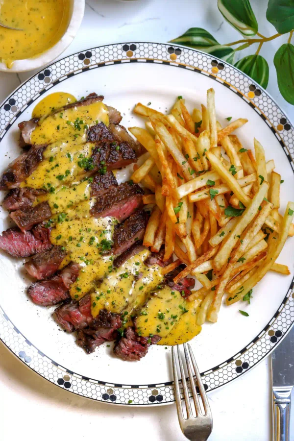 Top-down view of a round white plate with a black and gold geometric border holding sliced steak with sauce and a mound of french fries.