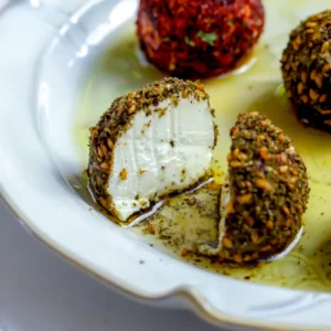 A close-up shot of a labneh ball coated in za'atar sliced in half to show the stark white, dense, and creamy yogurt cheese interior on a white plate with olive oil.