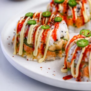 Three mini sushi bundt cakes on a white plate drizzled with Japanese mayo and sriracha, topped with serrano slices and shichimi togarashi, with spicy salmon and cucumber layers visible.