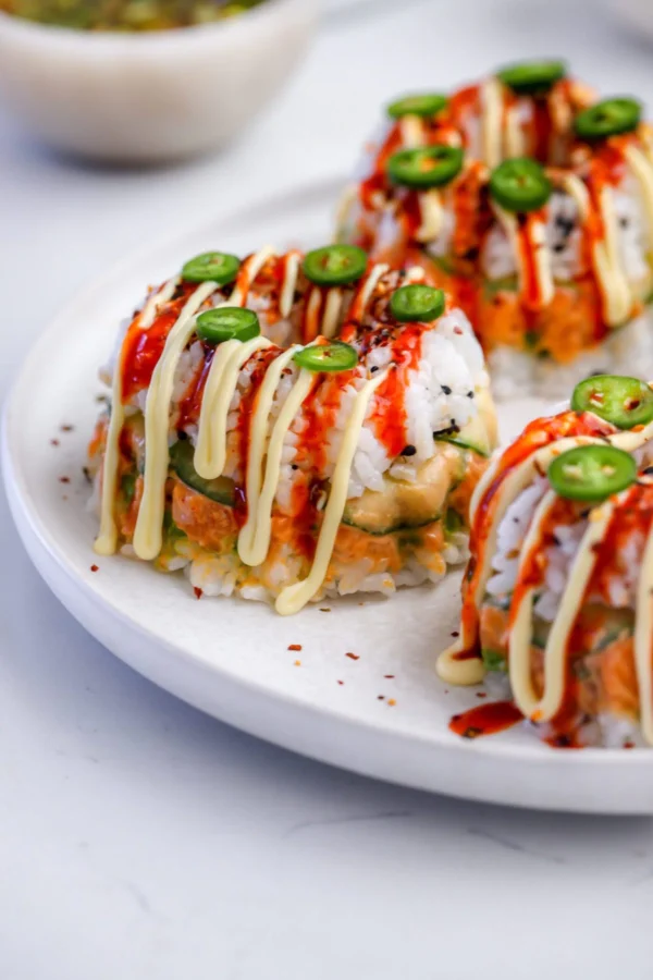 Three mini sushi bundt cakes on a white plate drizzled with Japanese mayo and sriracha, topped with serrano slices and shichimi togarashi, with spicy salmon and cucumber layers visible.