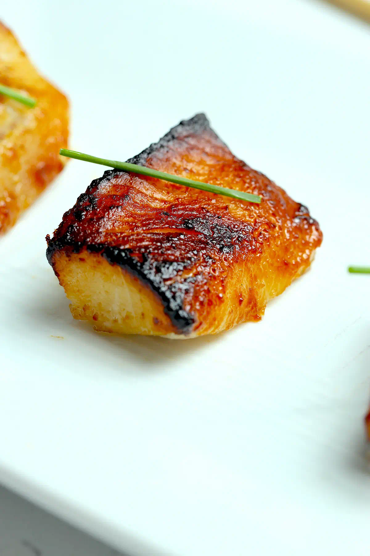Close-up of miso black cod, Nobu-style, with a caramelized amber crust and charred edges, garnished with a chive, the flaky white interior showing at the cut edge.