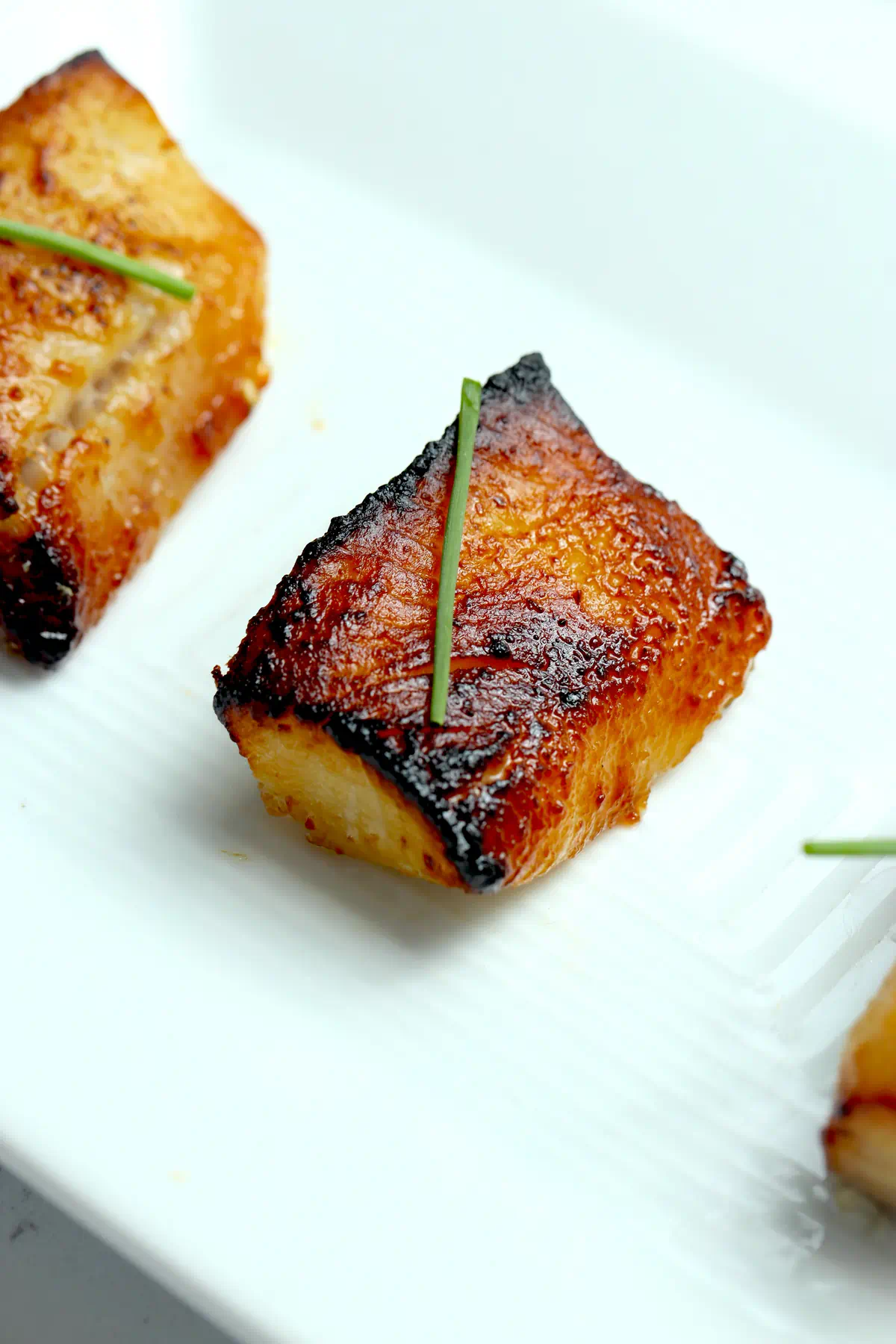Two pieces of Nobu-style miso black cod on a white platter, the front one topped with a chive, showing the caramelized crust and dark charred edges.