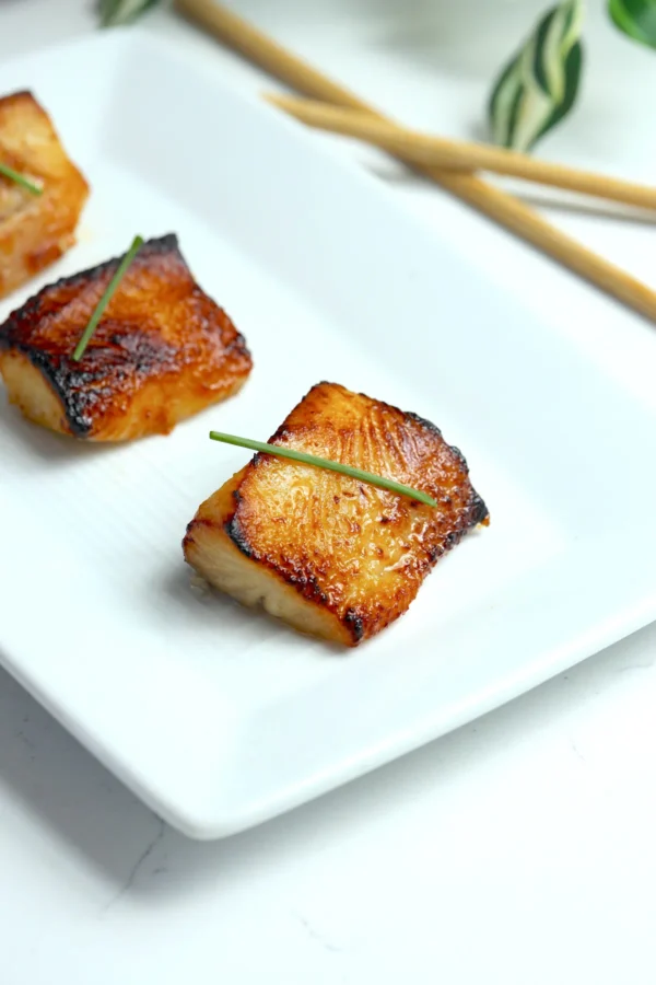 Three pieces of miso black cod, Nobu-style, arranged on a white platter with chopsticks, each garnished with a chive over the caramelized glaze.