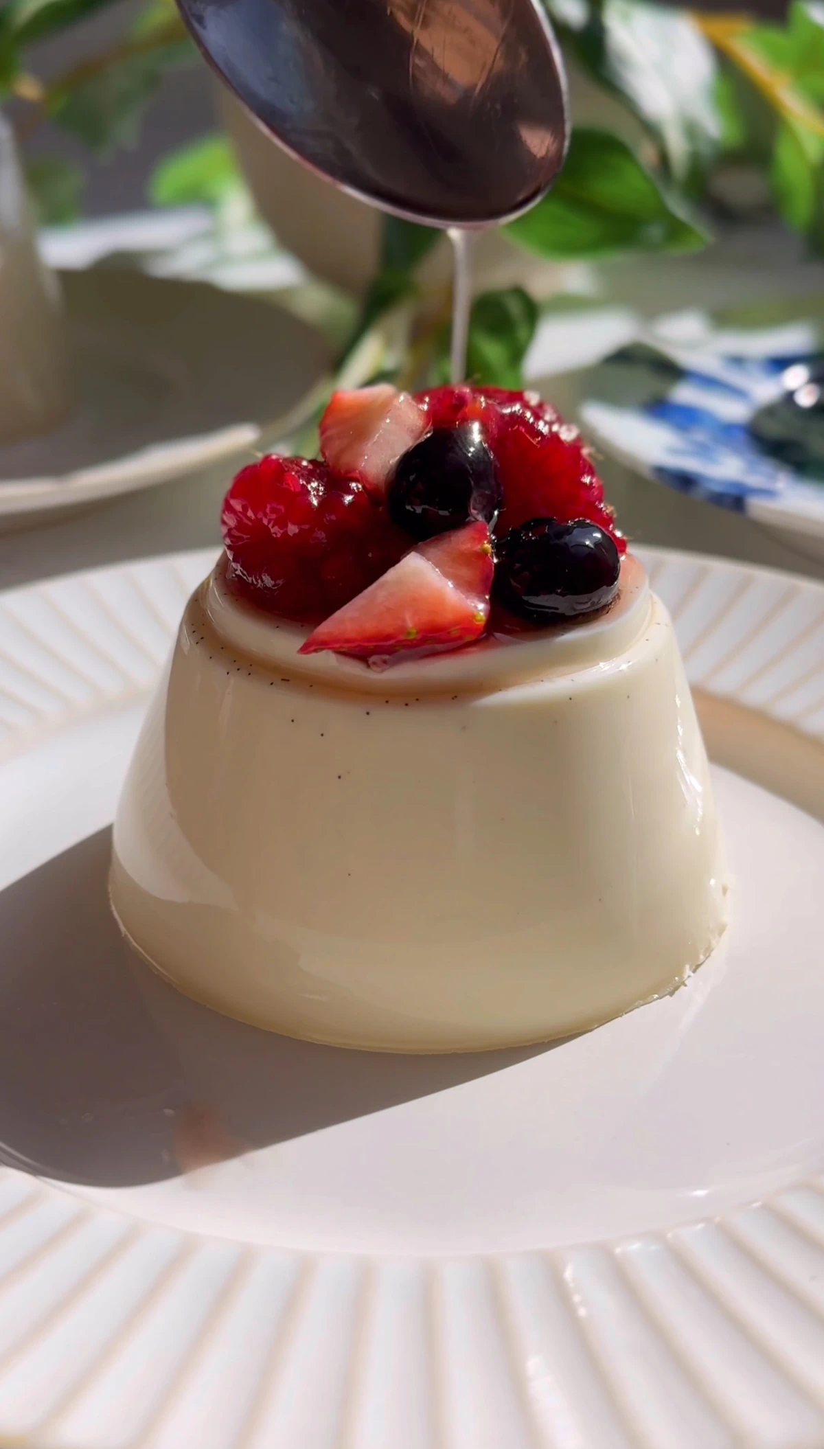 Close-up of berry panna cotta topped with raspberries, blueberries, and strawberries as syrup is spooned over.