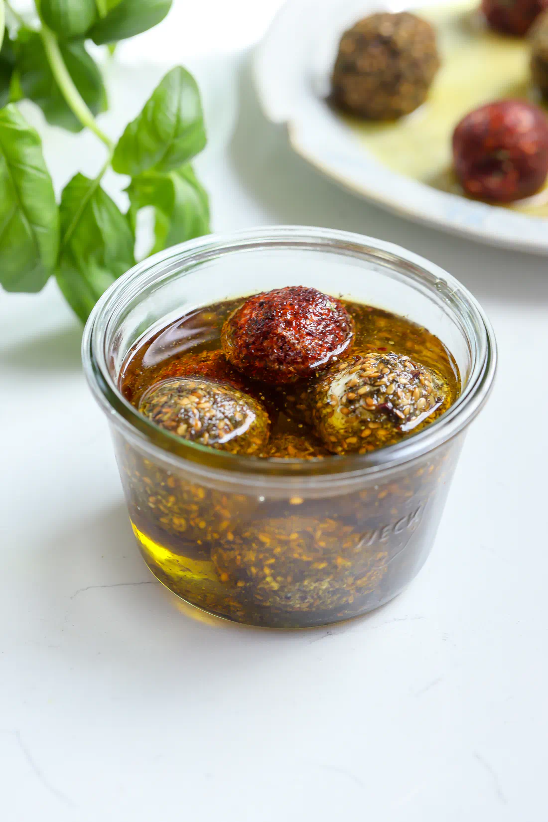 Top-down view of four labneh balls coated in red sumac and seedy za'atar, submerged in a glass jar of golden olive oil.