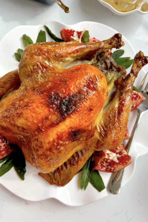Whole roasted Thanksgiving turkey with golden brown skin on a white scalloped platter with pomegranate wedges and sage leaves