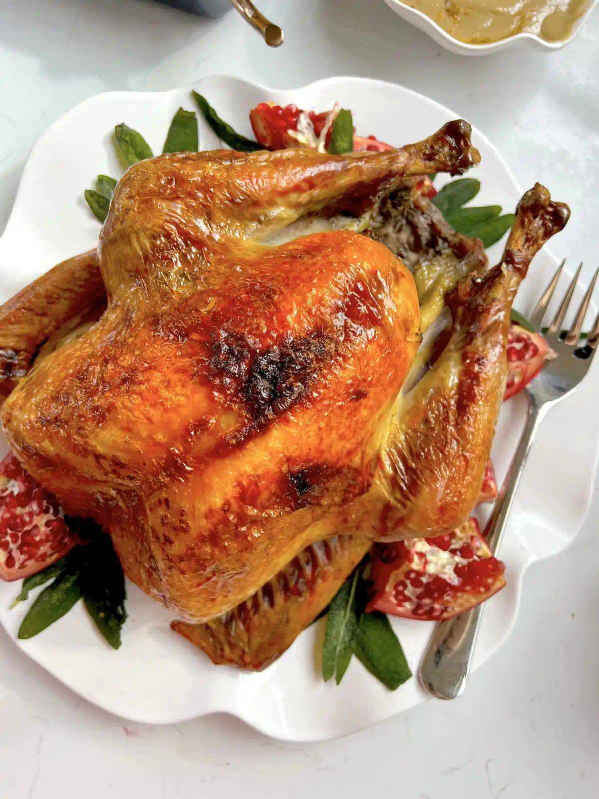 Whole roasted Thanksgiving turkey with golden brown skin on a white scalloped platter with pomegranate wedges and sage leaves