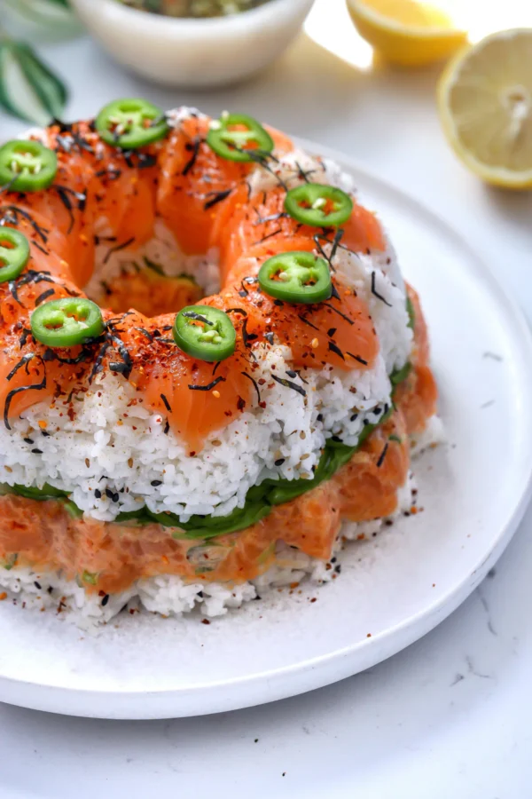 A full spicy salmon sushi bundt cake topped with glossy raw salmon slices, vibrant jalape&ntilde;os, and shichimi togarashi over sticky Calrose rice.