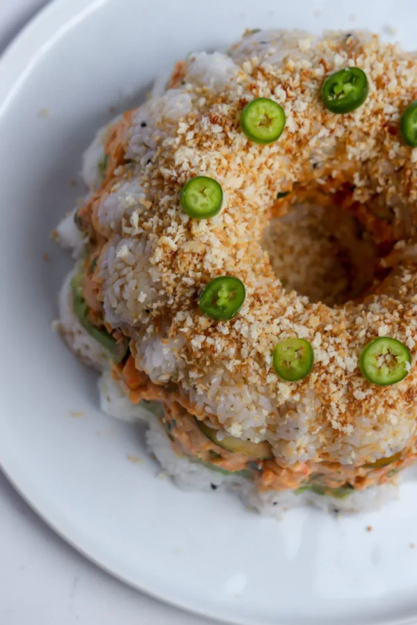 Overhead of shrimp sushi bundt cake topped with toasted panko, serrano slices, and unagi sauce.