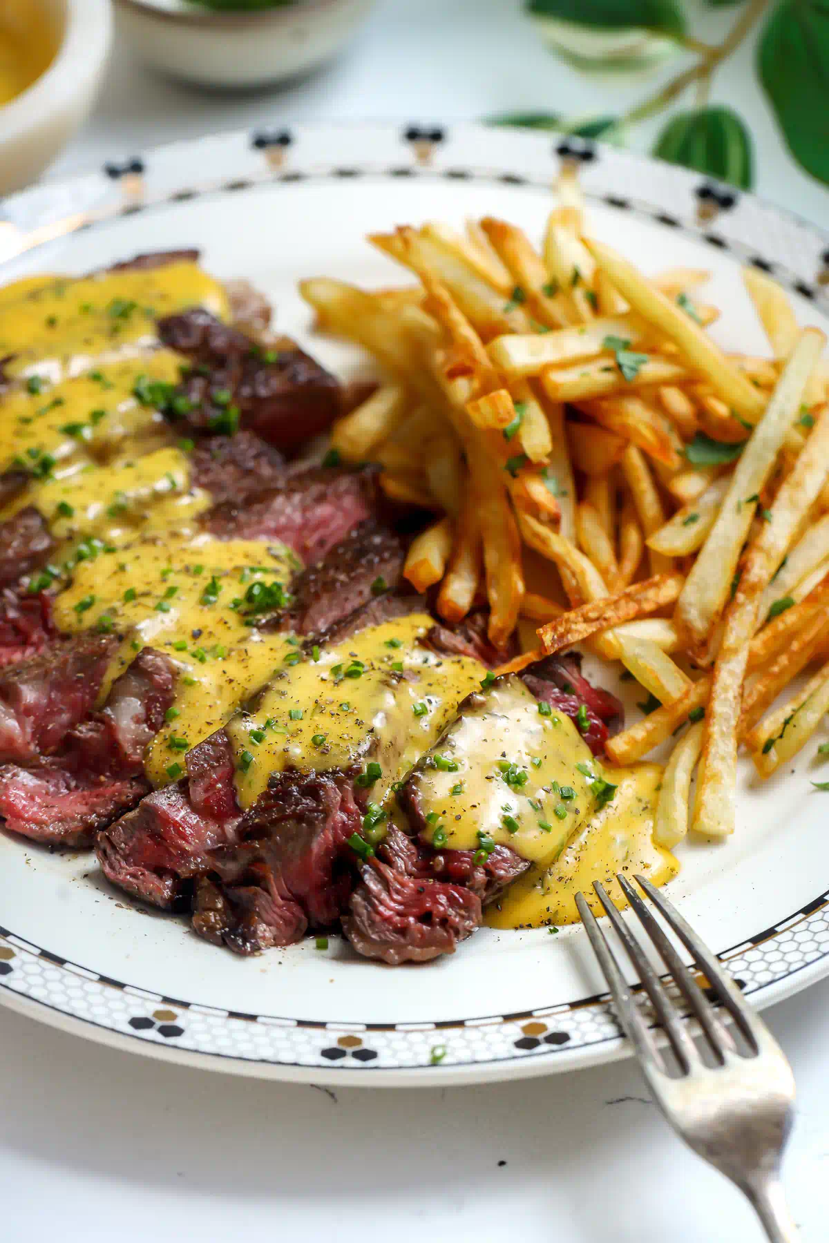 Close up of sliced steak drizzled with herb bearnaise sauce next to a pile of thin crispy frites with a silver fork in view.