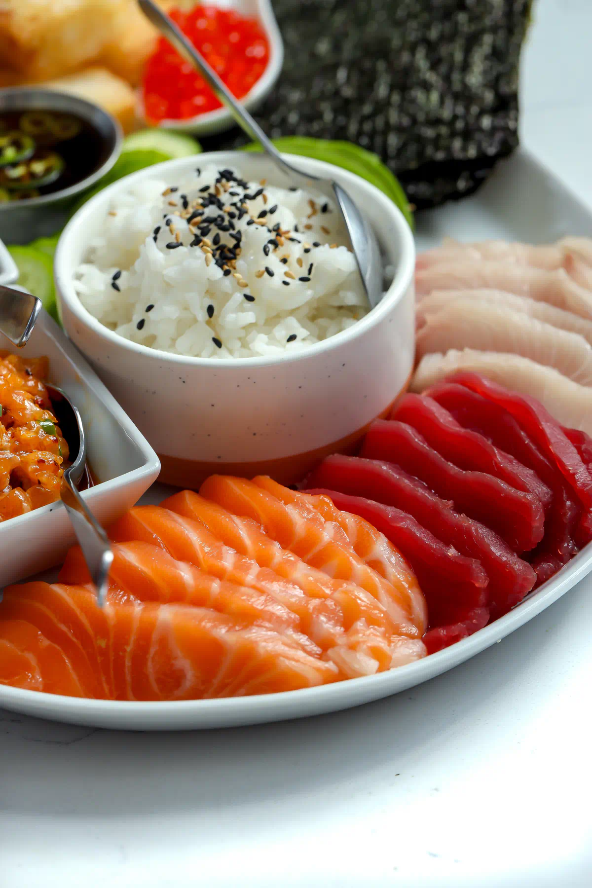 Salmon, tuna, and yellowtail sashimi fanned beside a bowl of sushi rice topped with black and white sesame seeds on a sushi board.