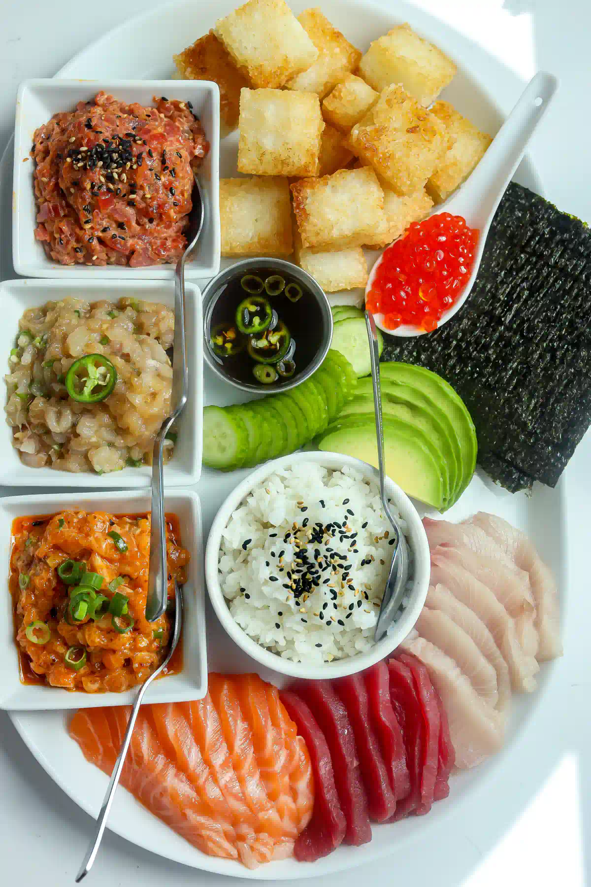 Full sushi charcuterie board from above with fried sushi rice cubes, spicy tuna, salsa macha salmon, yuzu jalapeño yellowtail, jalapeño ponzu, sushi rice, and sliced sashimi.