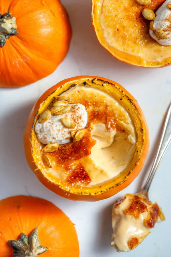 Overhead of pumpkin panna cotta in a hollowed mini pumpkin, brûléed sugar top cracked open, cinnamon whipped cream and roasted pumpkin seeds, whole pumpkins alongside.