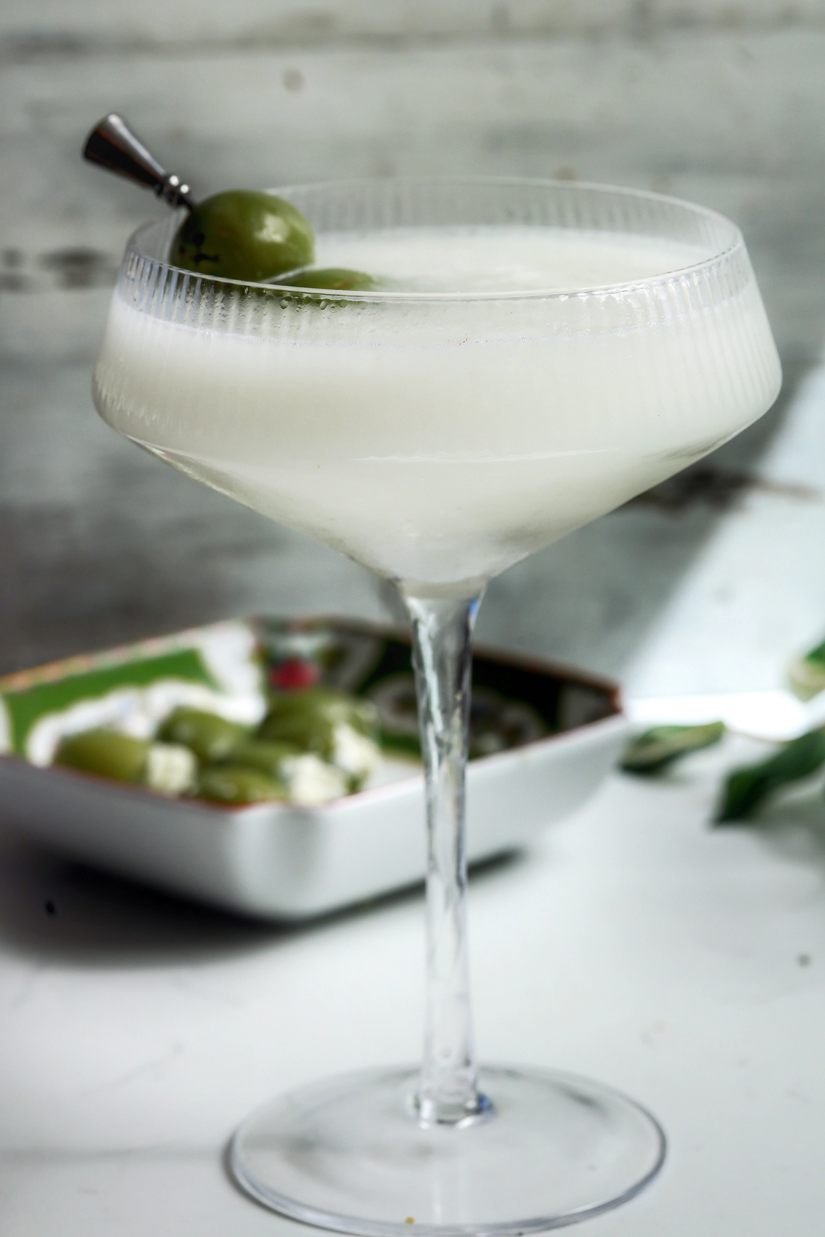 Milky blue cheese dirty martini in a ribbed coupe glass, garnished with two castelvetrano olives on a pick