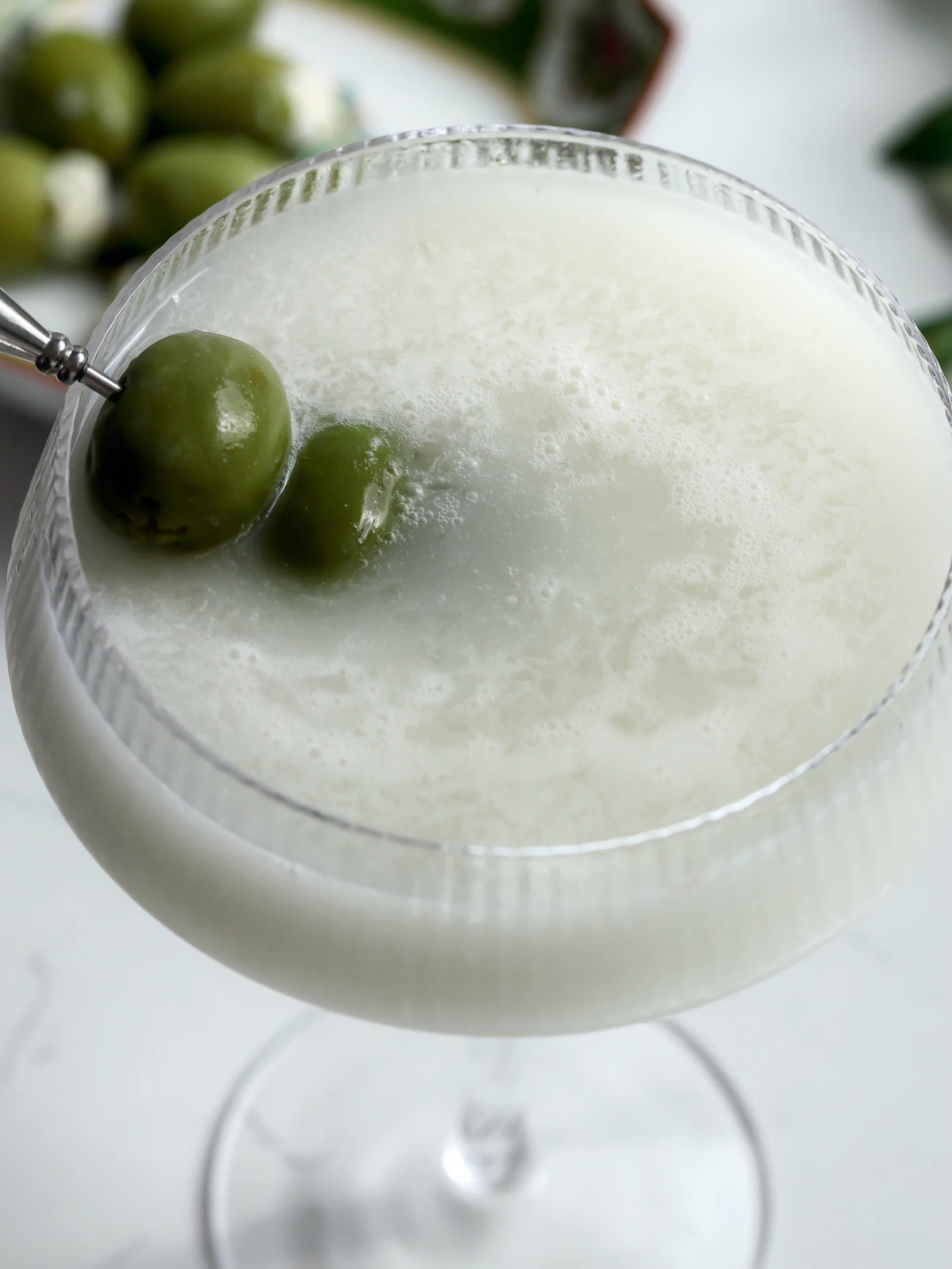 Overhead close-up of a blue cheese martini showing the milky, frothy surface and two green olives on a pick