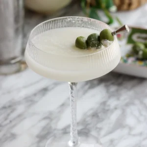 Blue cheese stuffed castelvetrano olives on a pick resting across a milky dirty martini, cocktail shaker behind