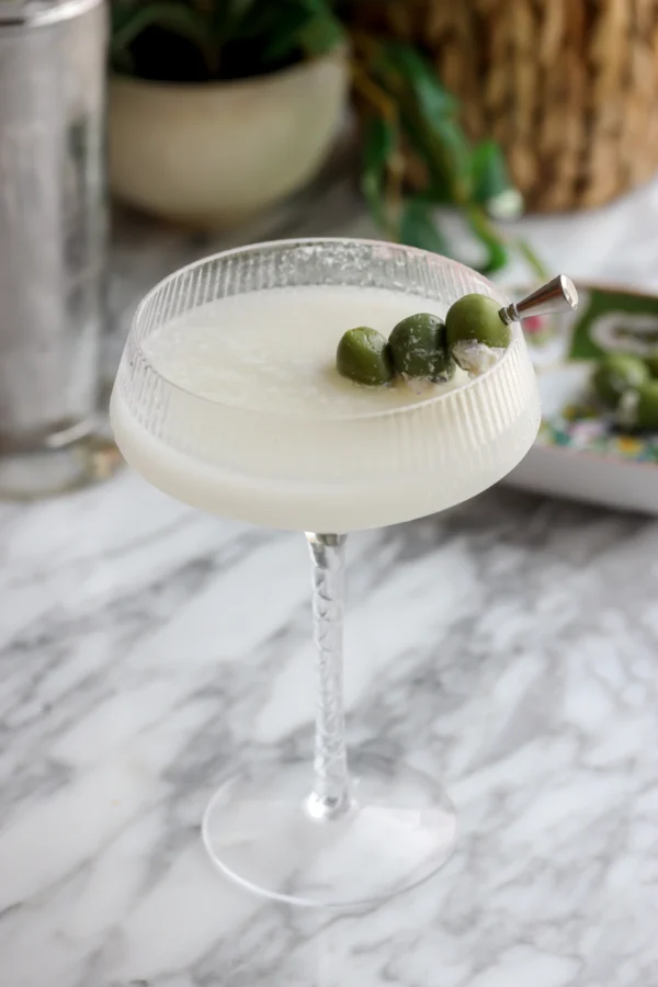 Blue cheese stuffed castelvetrano olives on a pick resting across a milky dirty martini, cocktail shaker behind