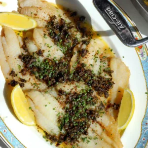 Overhead shot of freshly cooked petrale sole meuni&egrave;re garnished with chives and served with lemon wedges.