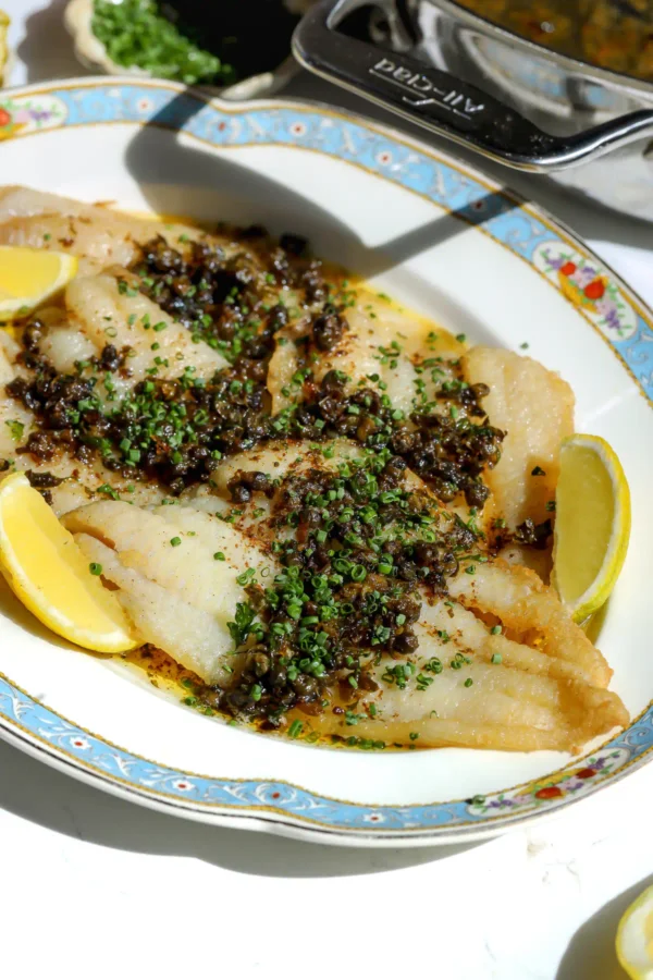 A close-up of a petrale sole meuni&egrave;re recipe featuring tender fish fillets topped with a browned butter and caper sauce on a pretty plate.