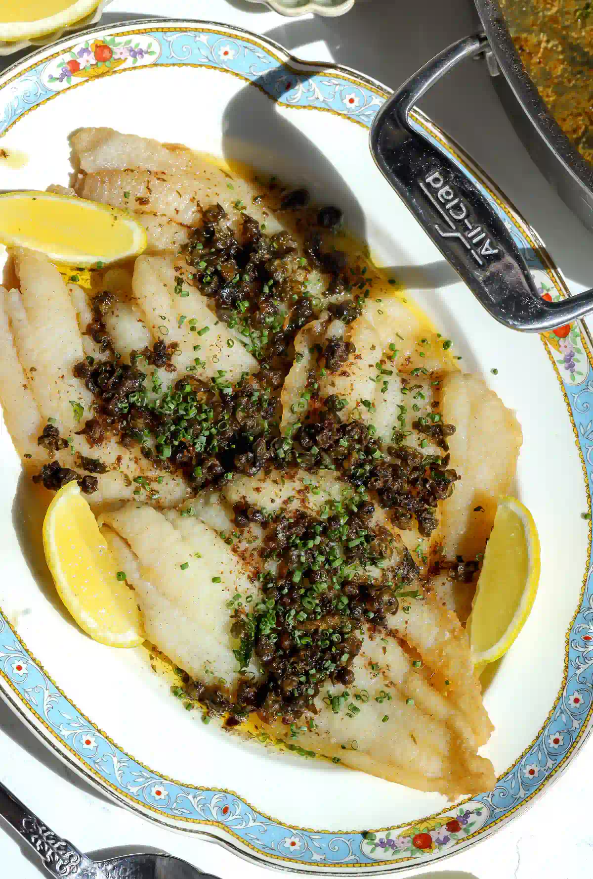 Overhead shot of freshly cooked petrale sole meuni&egrave;re garnished with chives and served with lemon wedges.