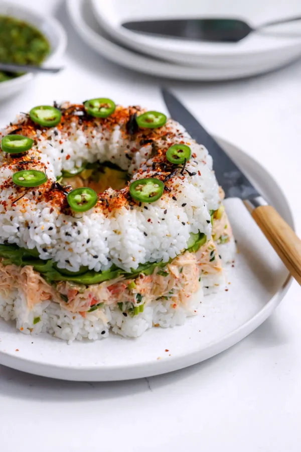 Overhead view of a sushi bundt cake recipe on a white platter topped with sliced serranos, shichimi togarashi, and black sesame seeds.
