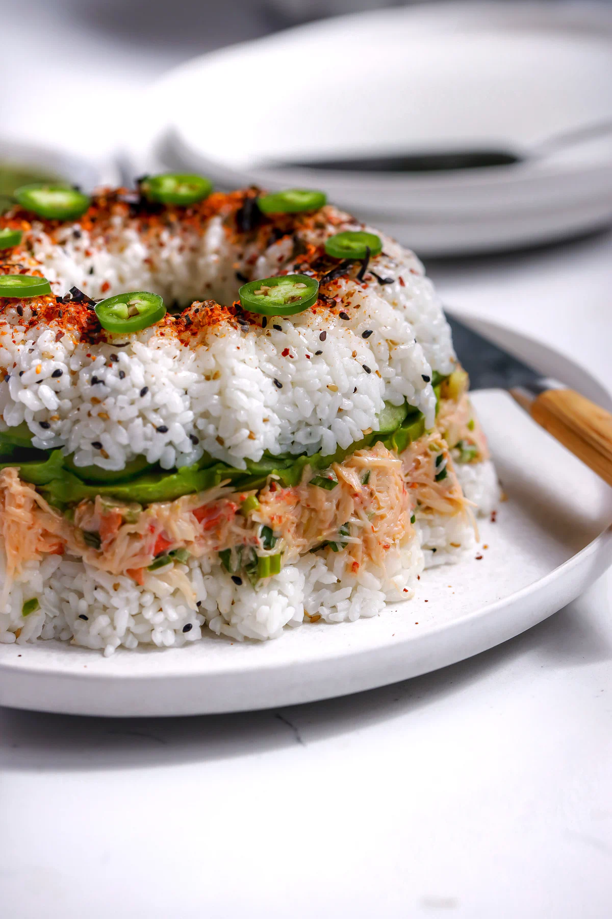 Profile view of a sushi ring appetizer showing distinct layers of seasoned rice, creamy crab tampico, sliced avocado, and crisp cucumbers.