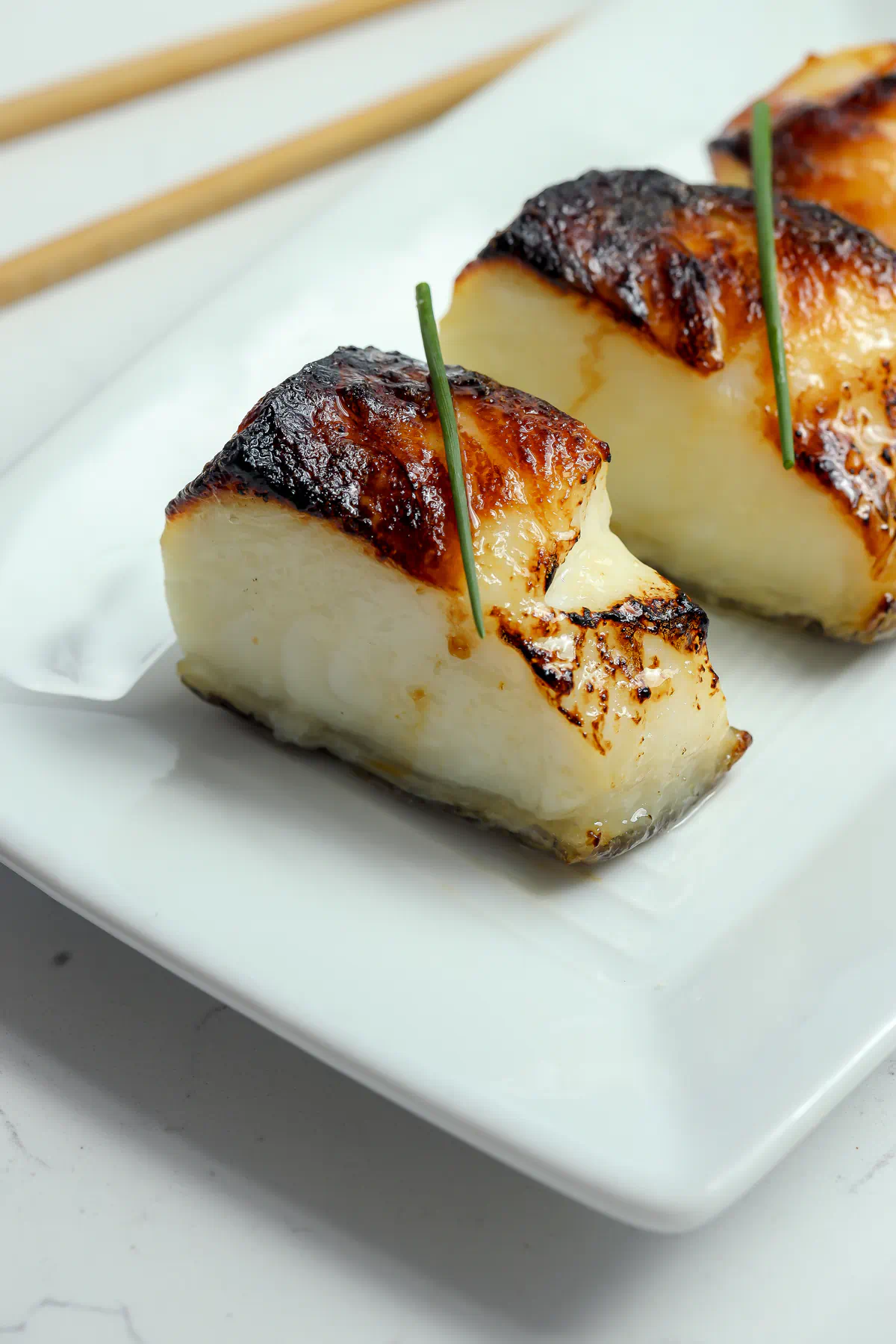Miso glazed Chilean sea bass fillets with deeply caramelized, charred tops and moist white flesh, each topped with a single chive, on a white plate