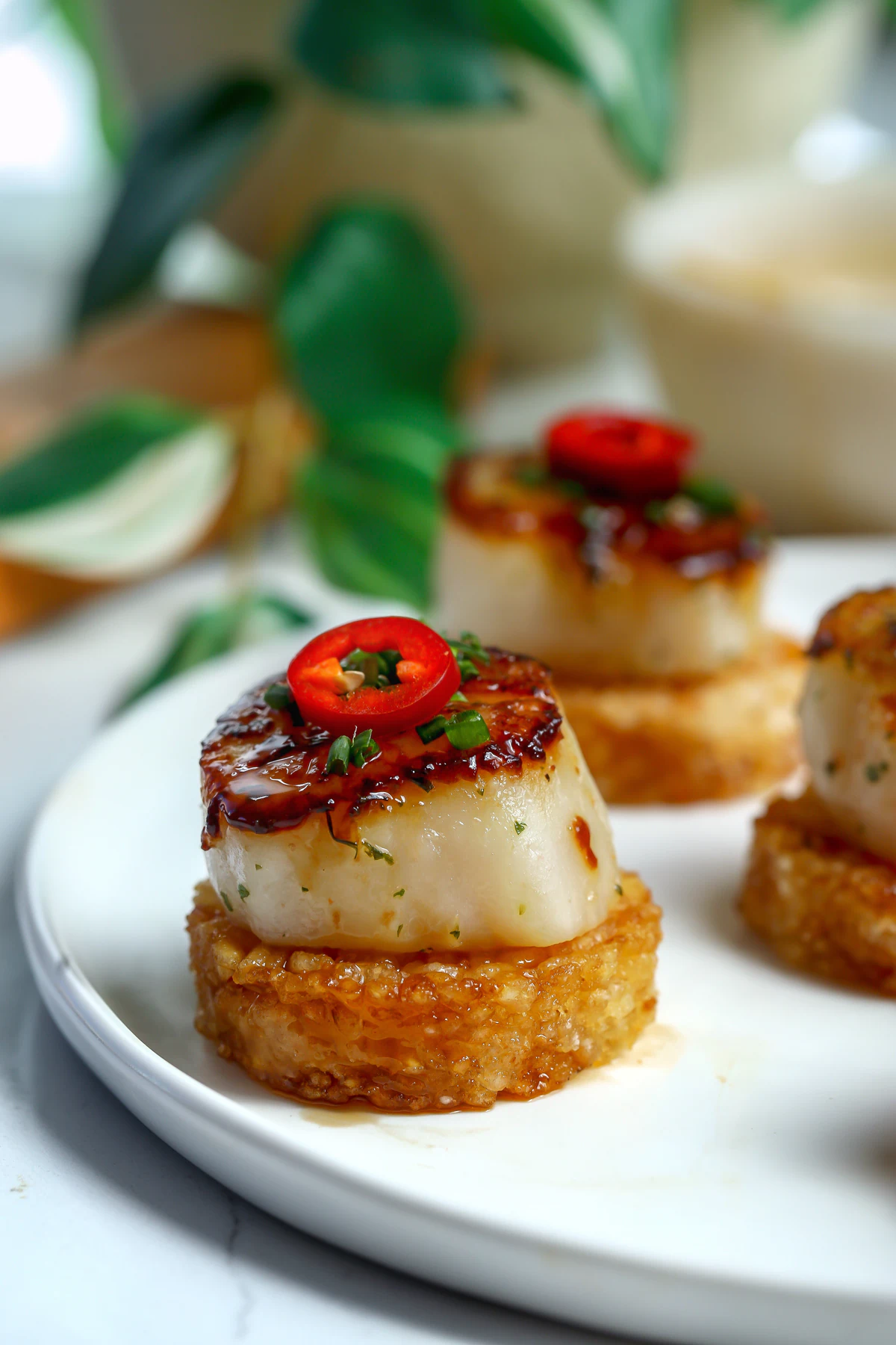 Golden-brown crispy rice rounds topped with a perfectly seared U-10 scallop, drizzled with a glossy sriracha agave glaze, and garnished with thin slices of Fresno chile and fresh chives on a light white ceramic plate.