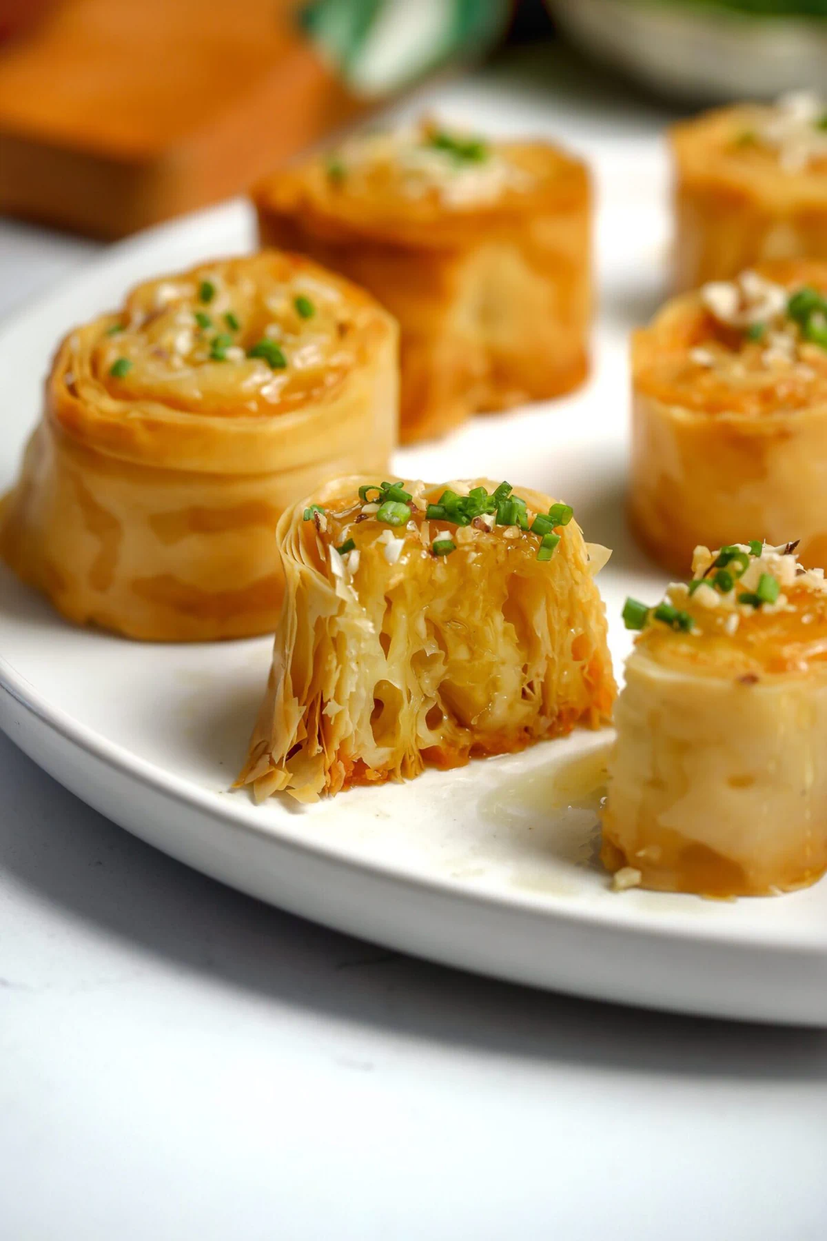 Bite-sized crispy baked phyllo with gruyere cheese between the layers cut in half to show the layers. Topped with honey, chives, and toasted almonds.