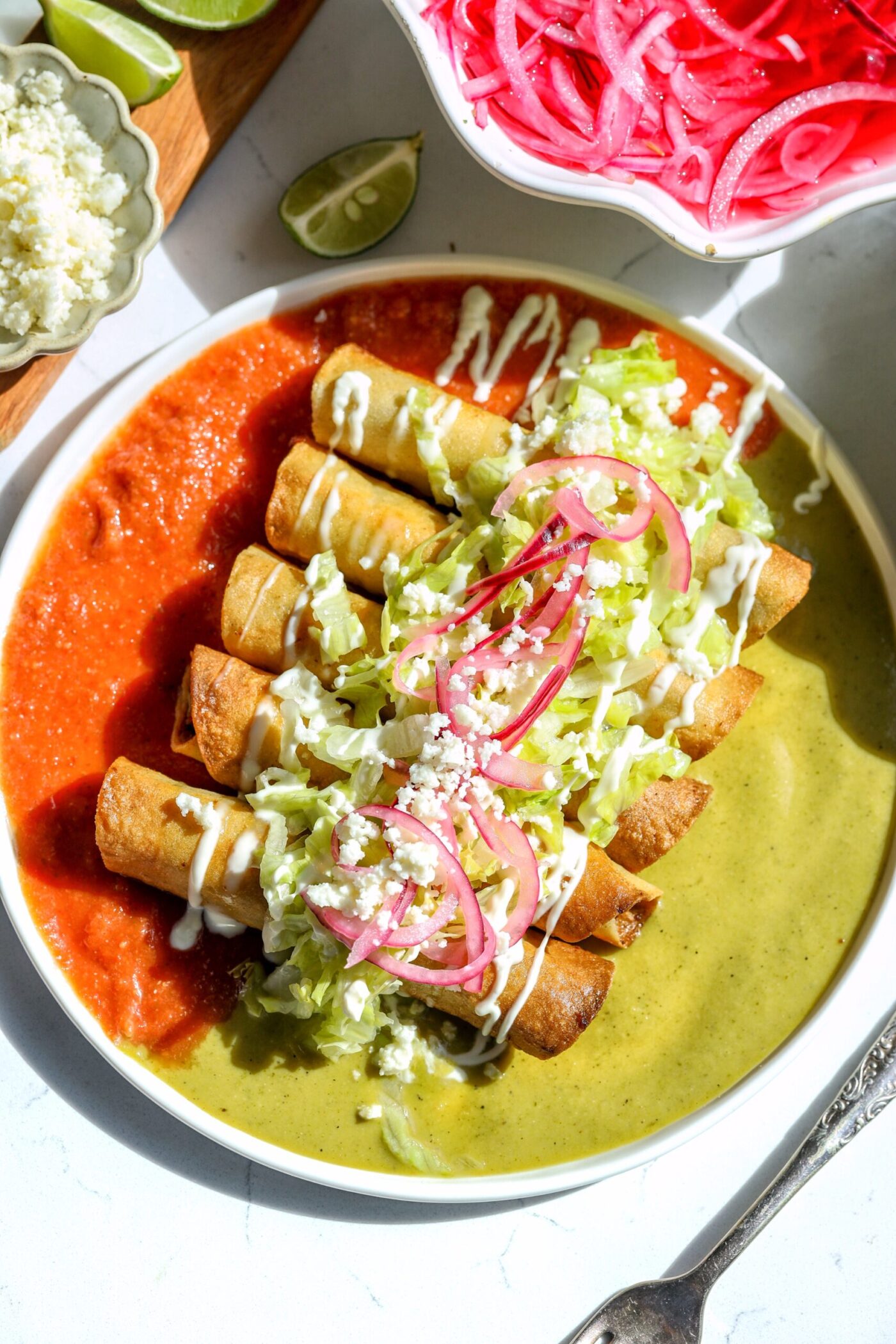 A plate of crispy flautas, garnished with shredded lettuce, pickled red onions, crumbled cheese, and drizzles of sour cream. The taquitos are divided by a dual sauce presentation: spicy red salsa on one side and creamy green salsa on the other.
