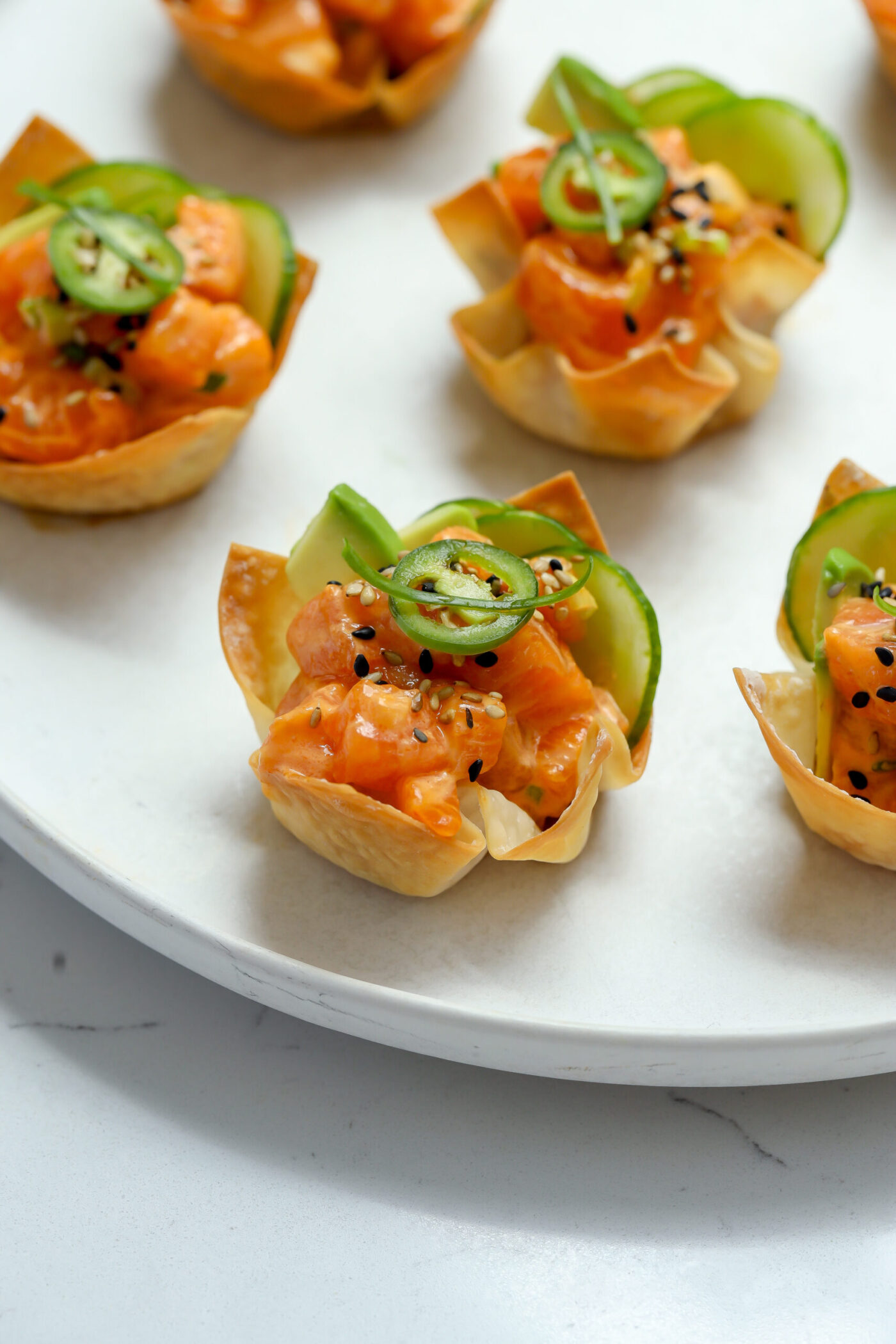 Close-up of a plate with Spicy Salmon Wontons featuring bite-sized appetizers in wonton cups, filled with a salmon mixture, topped with cucumber slices, jalapeño slices, and black sesame seeds.