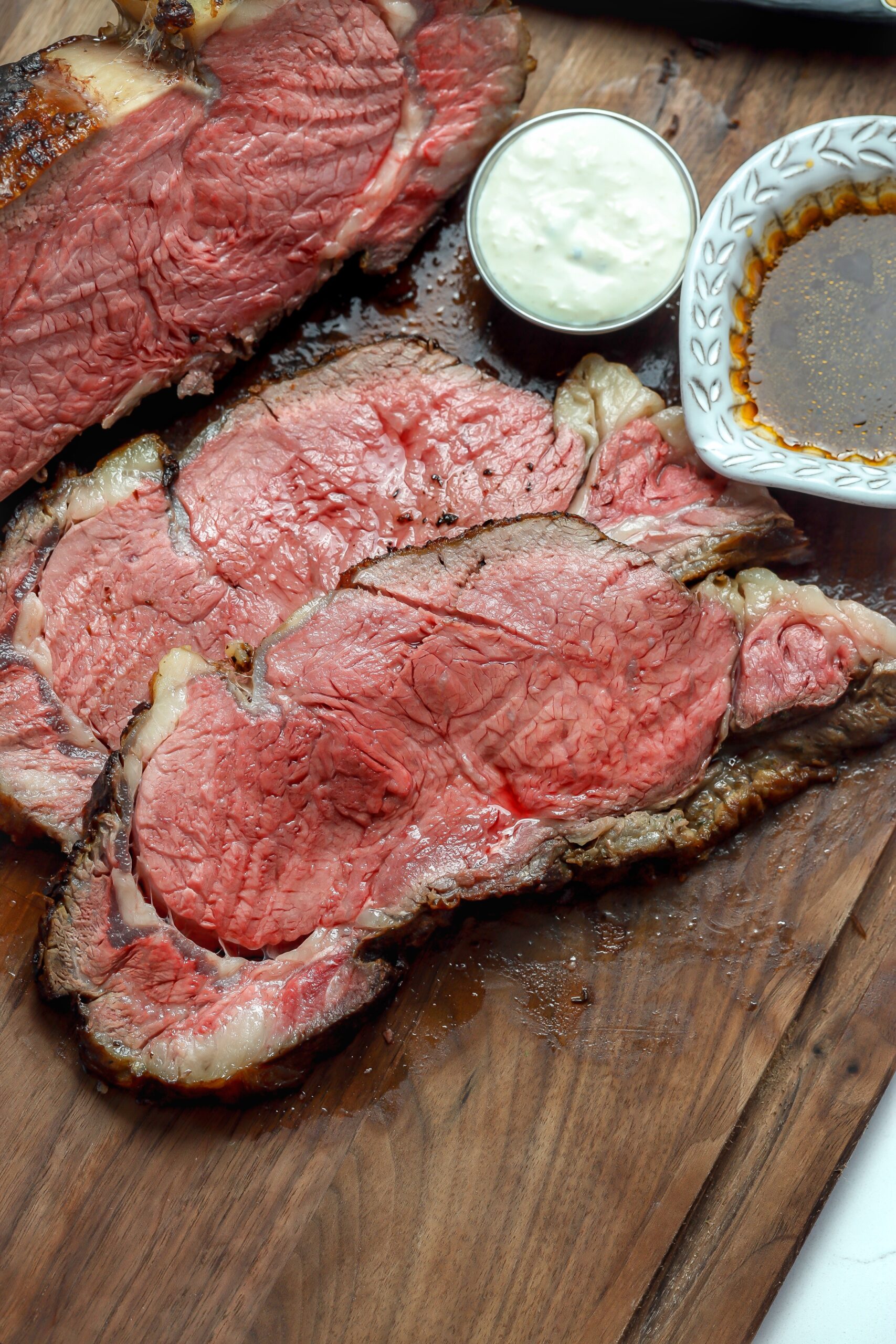 Perfectly seared and thinly sliced pieces of prime rib glistening on top of a dark wood cutting board. Next to it Is a ramekin of creamy horseradish nd a small gravy boat ramekin filled with savory au jus.
