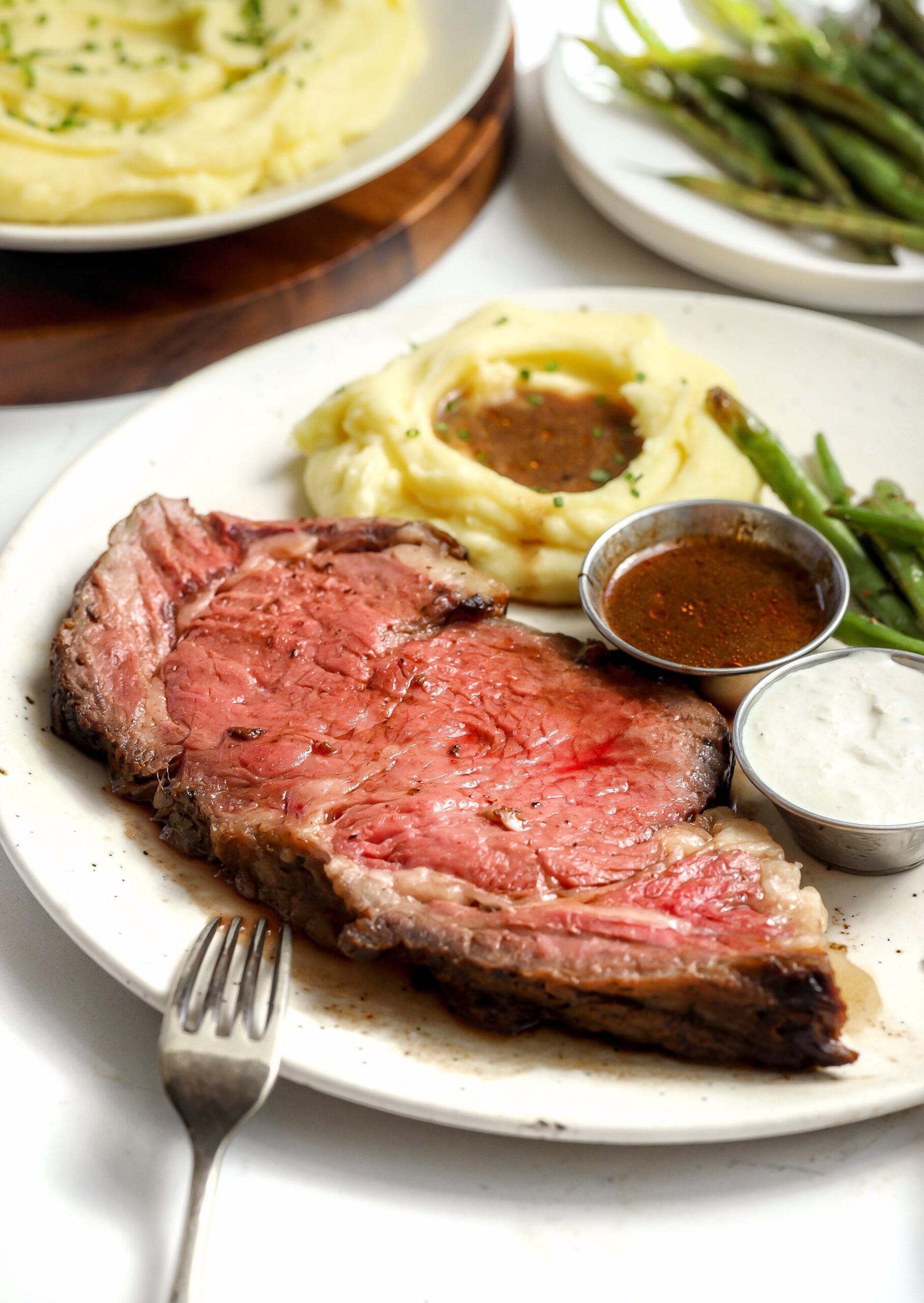 A plate with a juicy, medium-rare meat slice, surrounded by mashed potatoes with gravy and green beans. Additional sauces are served on the side. A fork rests beside the plate.
