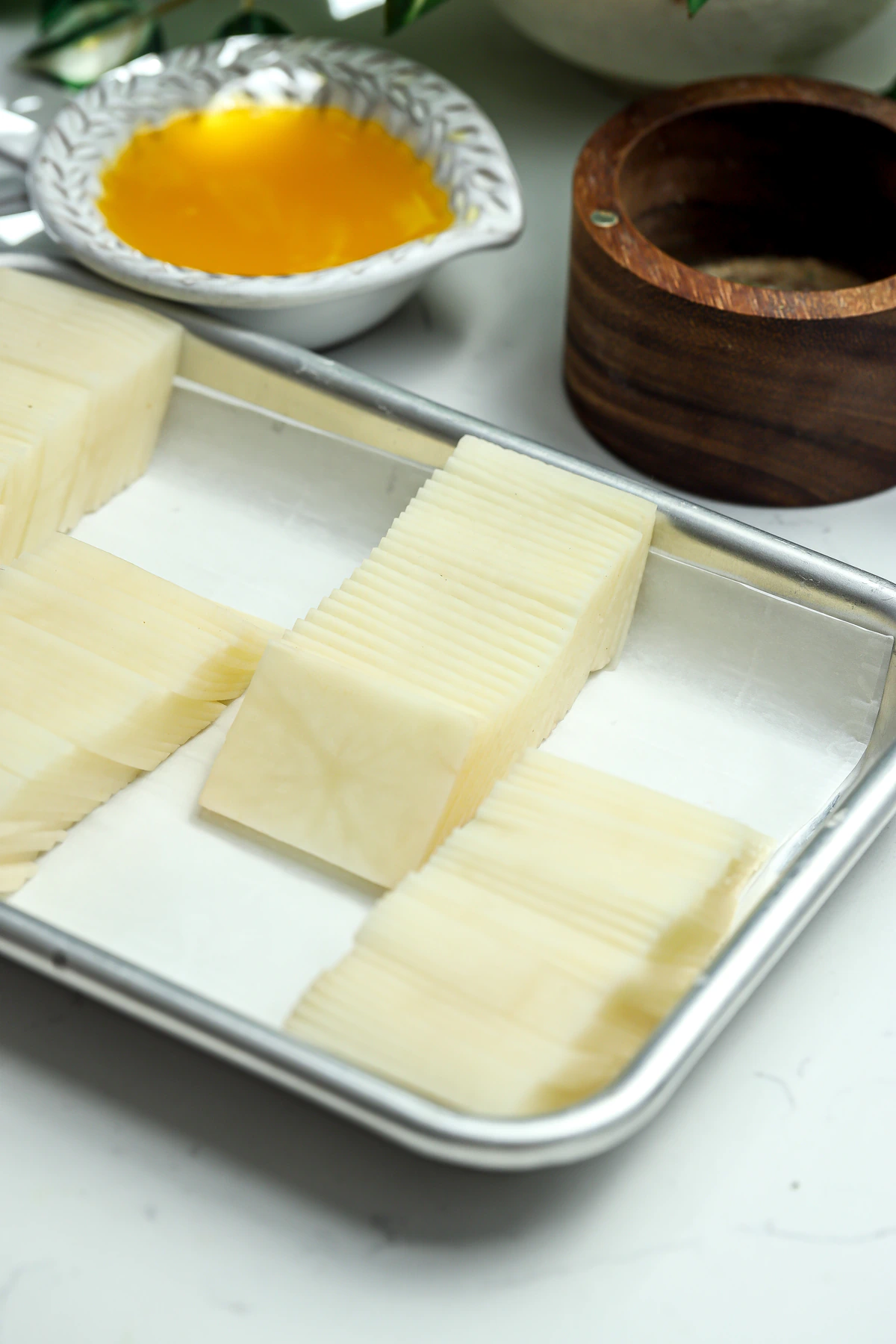 Thinly sliced potato stacks on a parchment-lined sheet pan next to a bowl of clarified butter and a wooden salt cellar ready to be baked