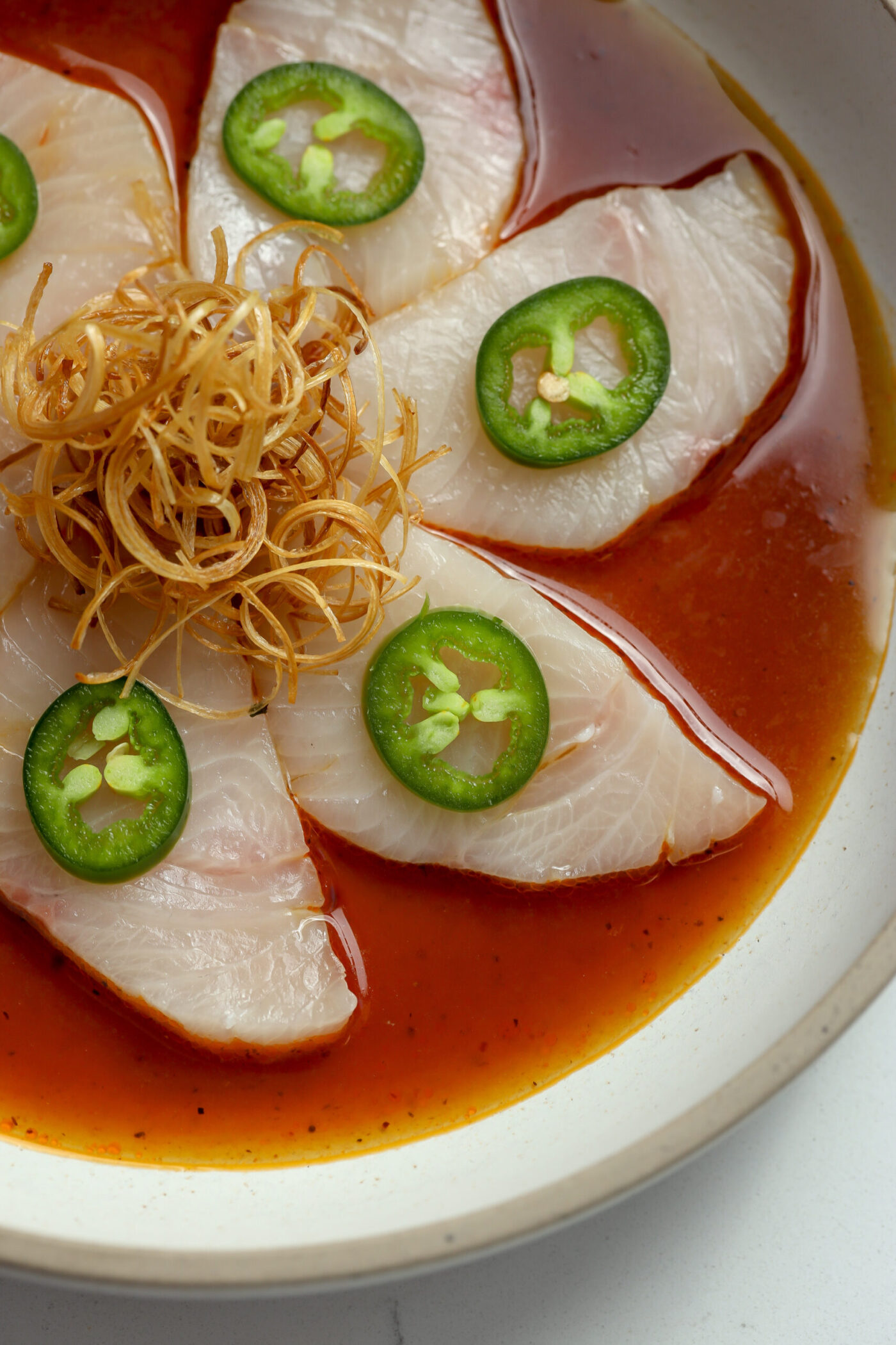 Thinly sliced hamachi resting in a delicious and spicy red sauce. Toppe with thin jalapeño slices and crispy, crunchy leeks in this crudo recipe.