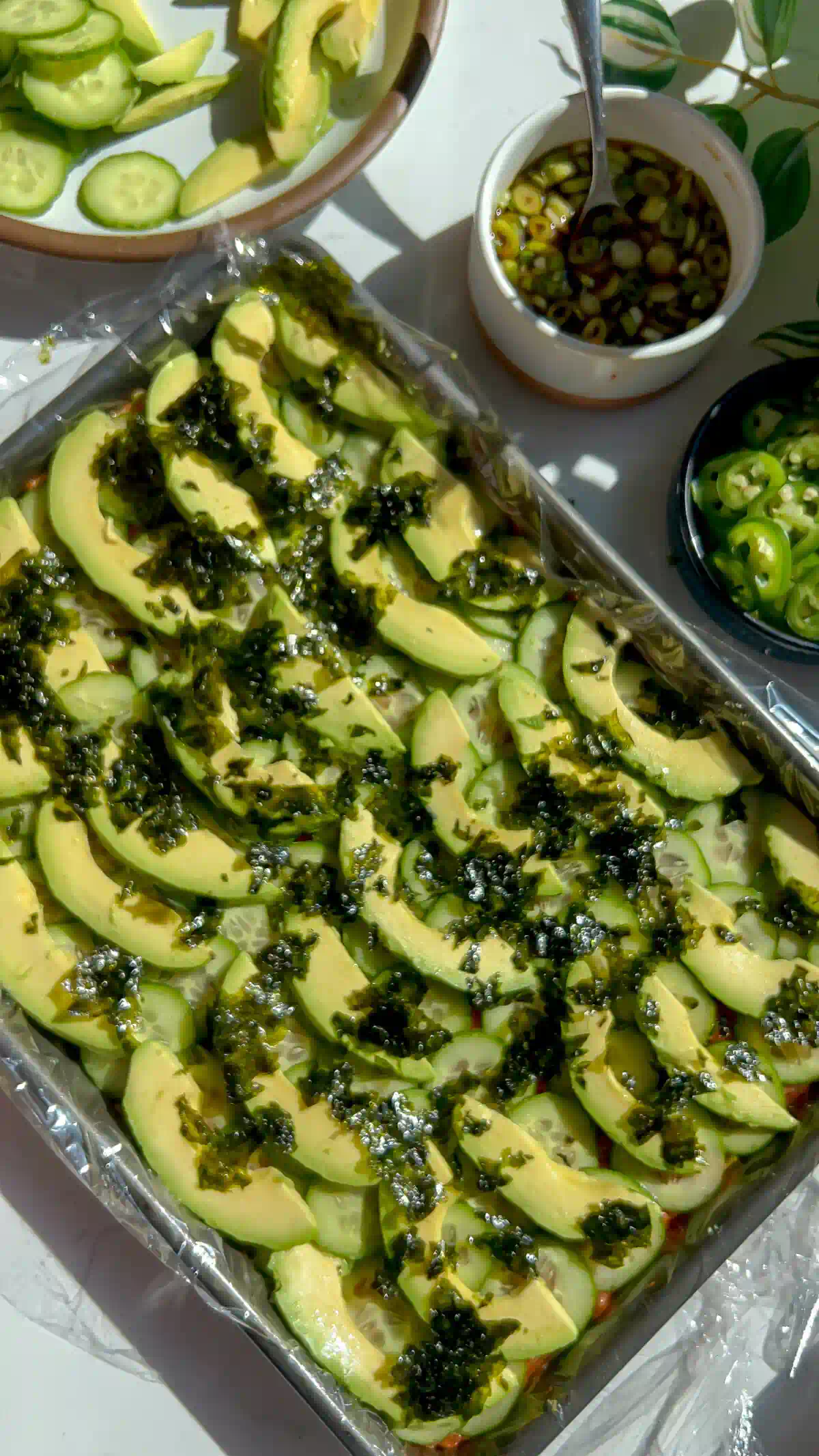 Avocado slices and crumbled nori layered over cucumber in a sheet pan of spicy tuna pressed sushi.