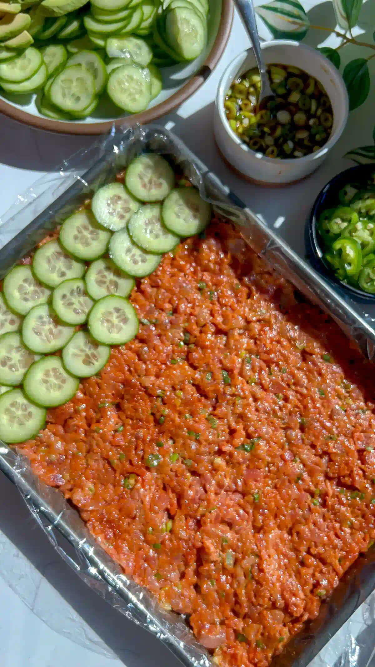 Cucumber rounds layered over gochujang spicy tuna in a plastic wrap lined sheet pan for pressed sushi.