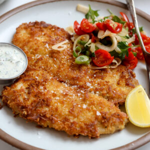 A potato and panko crusted and then pan fried petrale sole filet recipe with a tomato fennel salad, lemon, creamy tartare and fork on the plate next to it.
