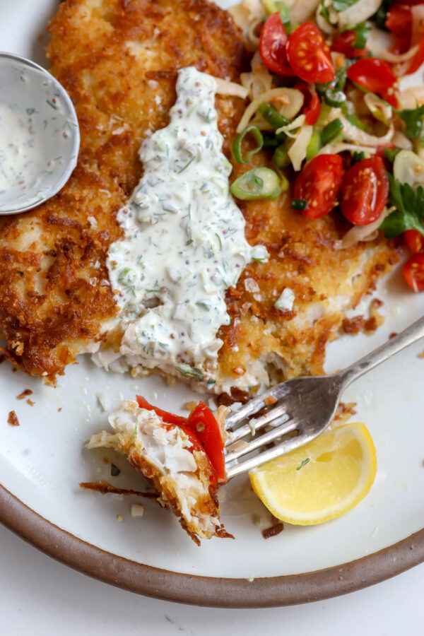 A potato and panko crusted and then pan fried petrale sole filet recipe topped with a tomato fennel salad and creamy tartare. the perfect bite of white fish and, salad and tartare is sitting on a fork next to a lemon wedge.