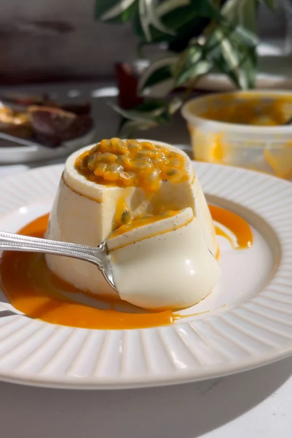 Spoon lifting a bite from a vanilla bean passionfruit panna cotta, passion fruit pulp on top and caramel pooled around the base.