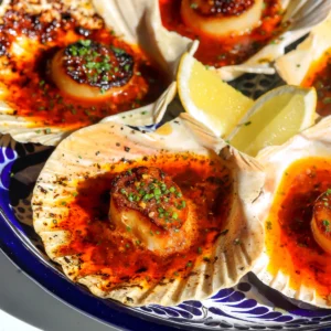 Overhead view of four salsa macha butter scallops broiled in shells on a blue Talavera platter with lemon wedges and chives.