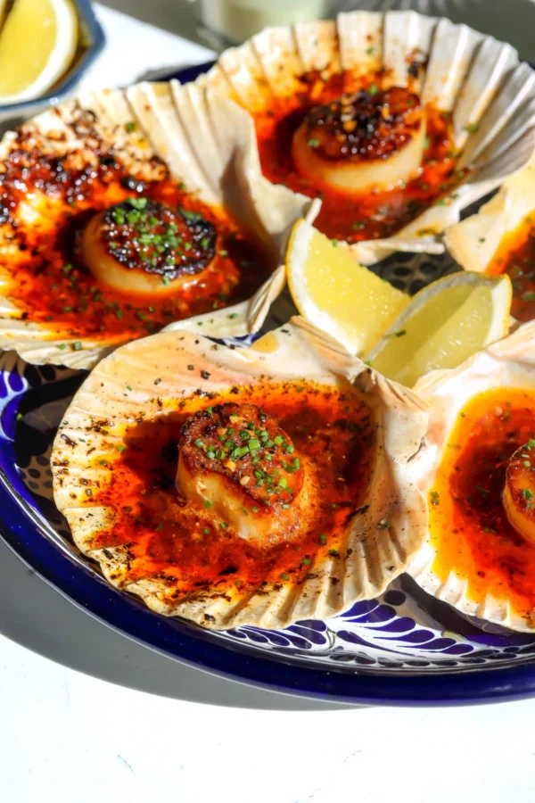 Overhead view of four salsa macha butter scallops broiled in shells on a blue Talavera platter with lemon wedges and chives.