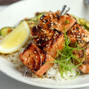 Miso salmon skewers over white rice, topped with sesame seeds and curled scallion, with a lemon wedge alongside.