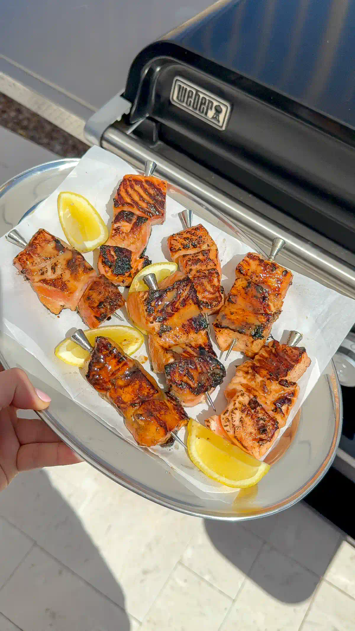 Miso salmon skewers with lemon wedges on a metal platter, held in front of a Weber griddle outdoors.