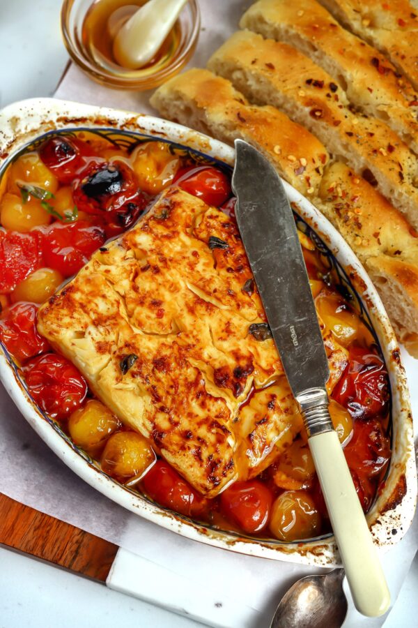 Baked Feta and Tomato in seasoned oil with garlic fresh out of the oven and golden with a spreading knife on top and seasoned bread next to it ready to dip.