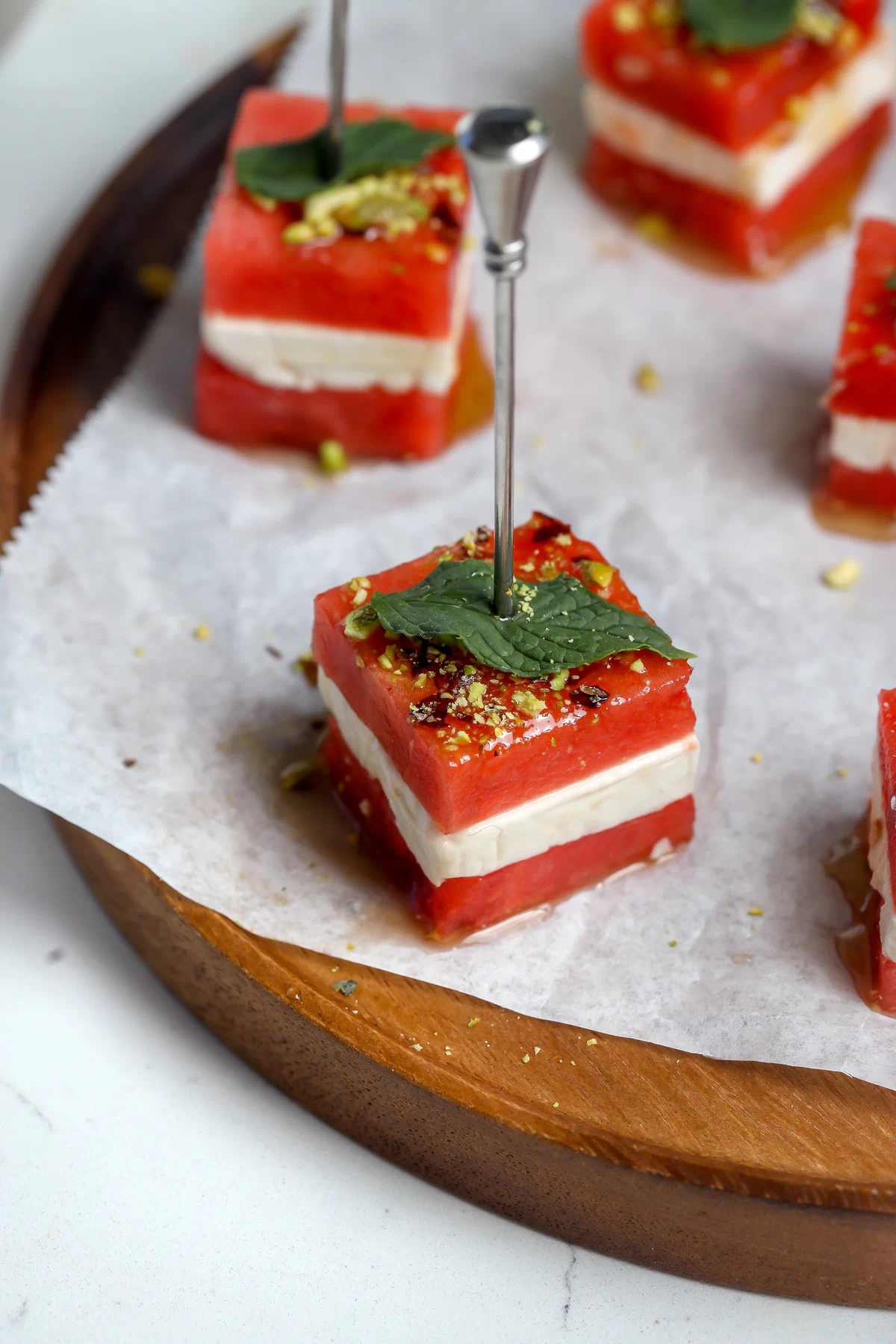 Watermelon feta appetizer bites on skewers, topped with hot honey balsamic glaze, crushed pistachios, and fresh mint on a wood plate.